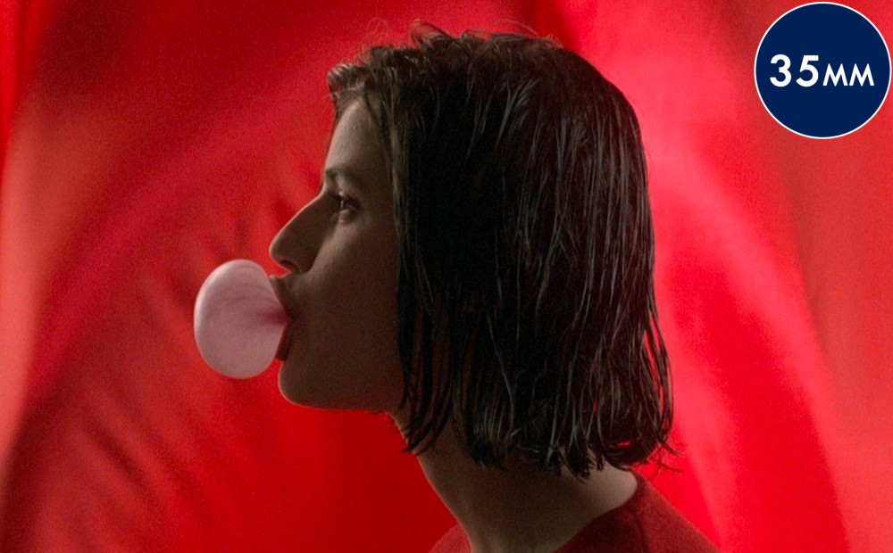 Actor Irene Jacob in profile against a red background, blowing a bubble with chewing gum.