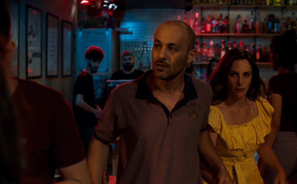 A man holds the hand of a woman in a yellow dress, leading her through a bar.