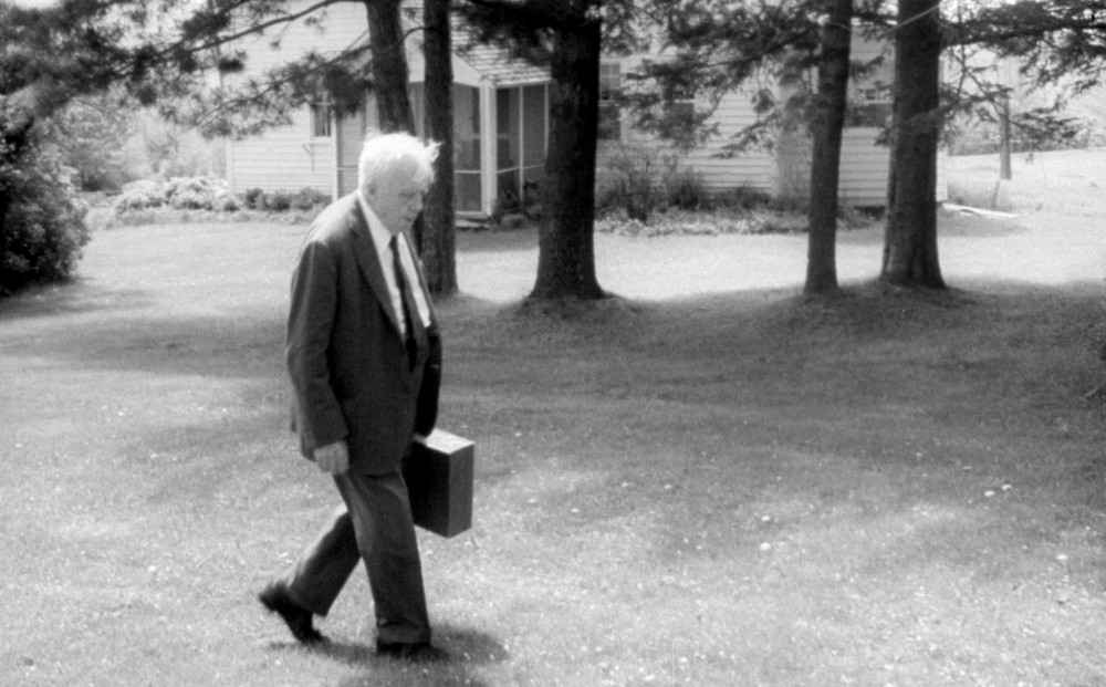 Robert Frost walks across his lawn, wearing a suit and holding a briefcase.