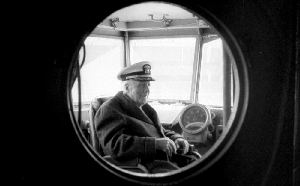 Robert Frost, as seen through a porthole into the captain's quarters on a ship; he wears a captain's hat.