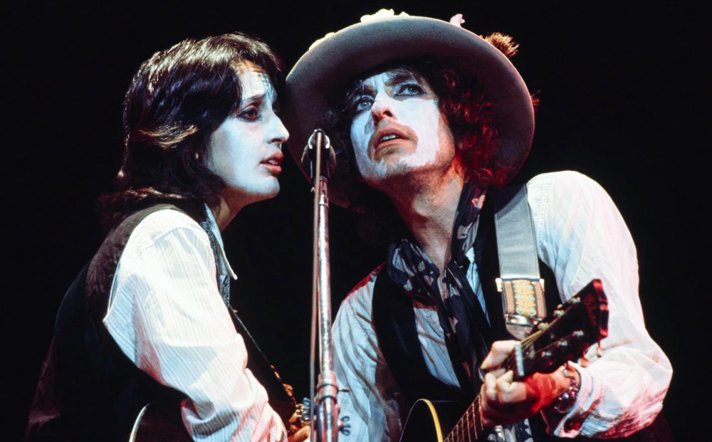 Joan Baez and Bob Dylan sing and play guitar together, sharing one microphone and wearing matching white face paint, white button-down shirts, and vests.
