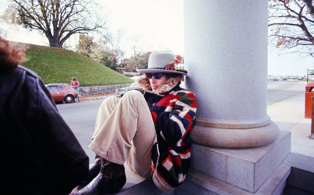 Bob Dylan sits against a marble column, huddled in a hat, sunglasses, cowboy boots, and a colorful coat.