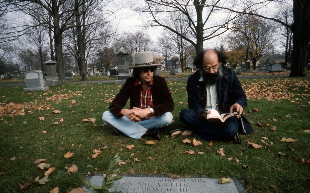 Bob Dylan and Allen Ginsberg sit together by a gravestone in a cemetery.