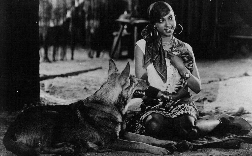 Actor Josephine Baker sits on the ground, smiling, next to a dog.
