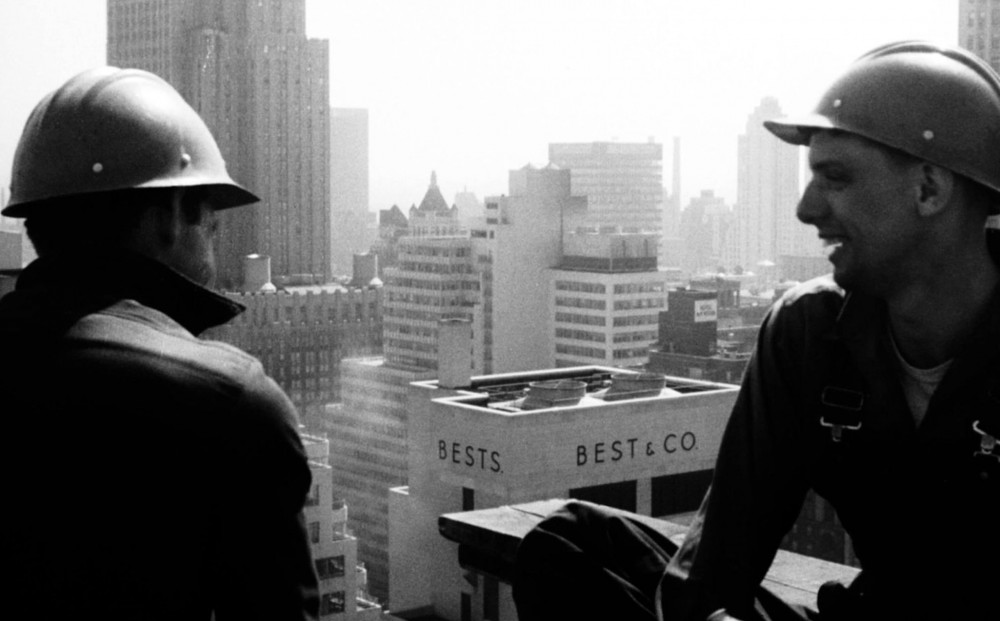 Two men wearing construction helmets sit on a rooftop.