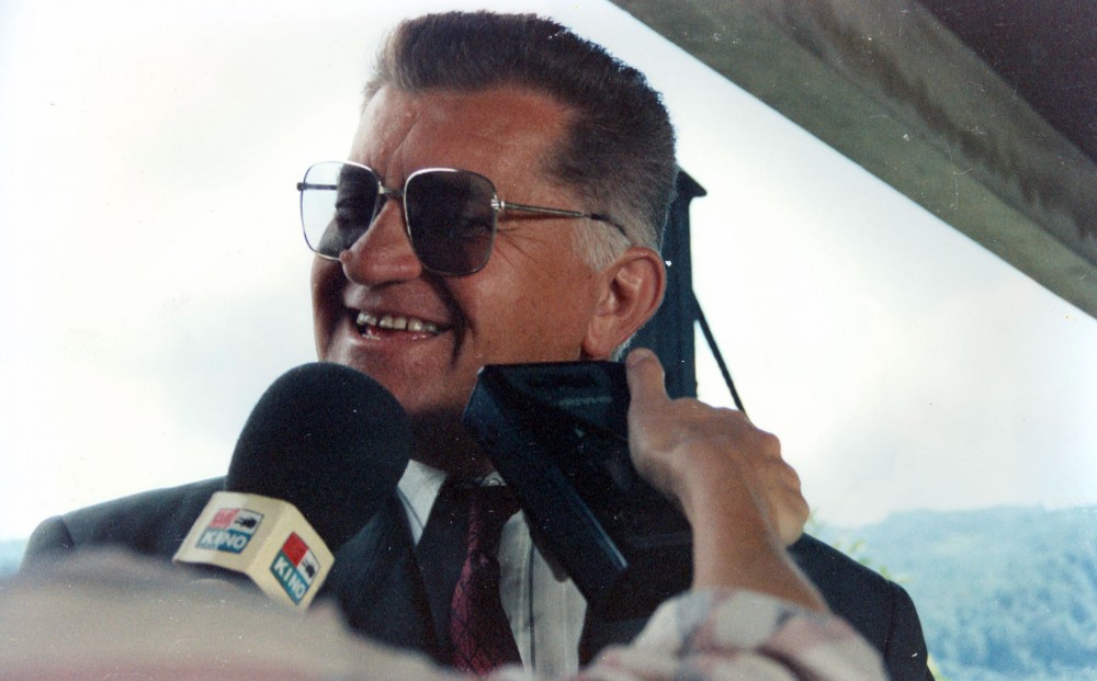 A man smiles in front of a microphone; someone holds a recording device up to his mouth.