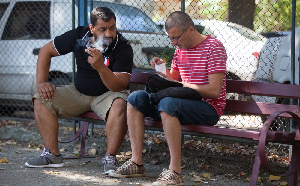 Two men sit next to each other on a bench in front of a chain-link fence; one looks at a piece of paper and the other smokes.