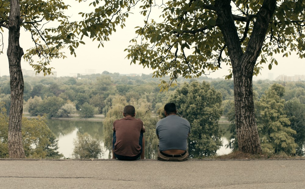 Two men sit along a river or lake, their backs to the camera.