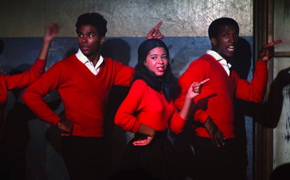 Two men and a woman stand in a line, wearing matching red sweaters and dancing.