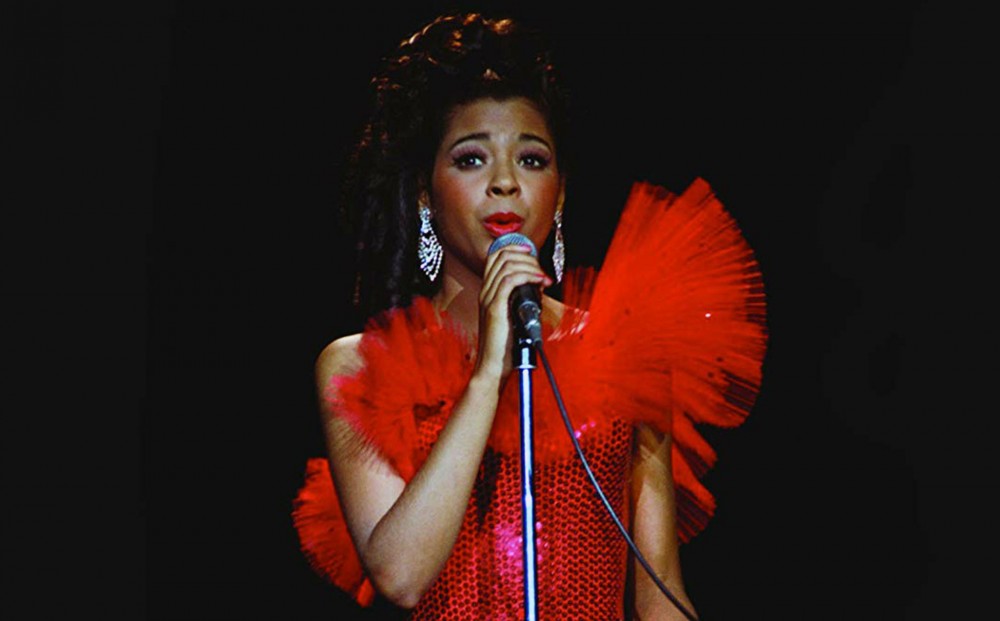 A woman wearing a red feather boa and red sequined dress holds a microphone and sings.