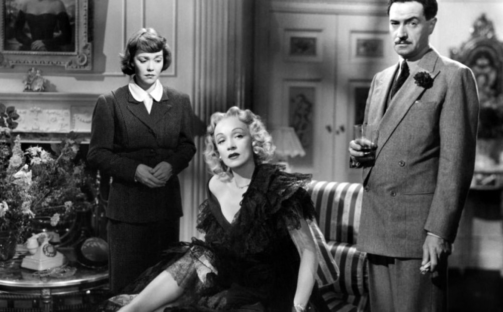 The main actors are in a sitting room, with Marlene Dietrich seated between them while they stand.