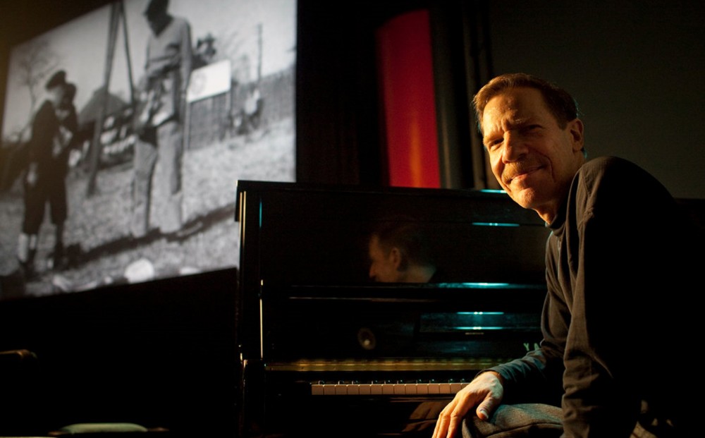 Piano accompanist Steve Sterner sits at a piano in the theater; a film plays on the screen in the background.