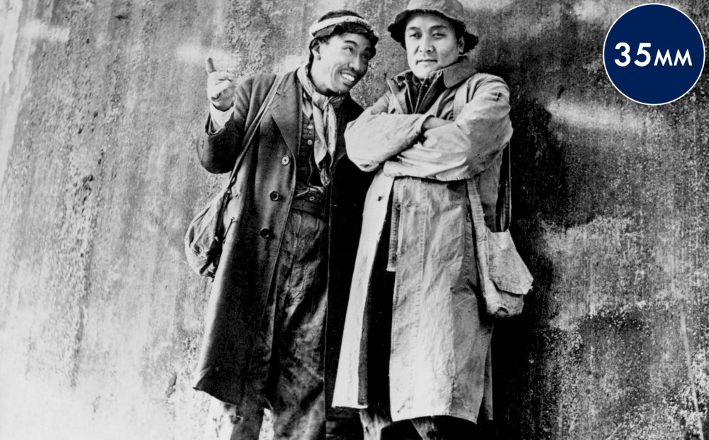 Two men in coats stand next to each other near a wall.