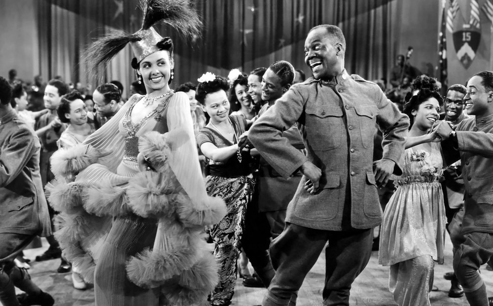 Bill Robinson and Lena Horne pose with their arms on their hips on the dance floor of a large nightclub. 