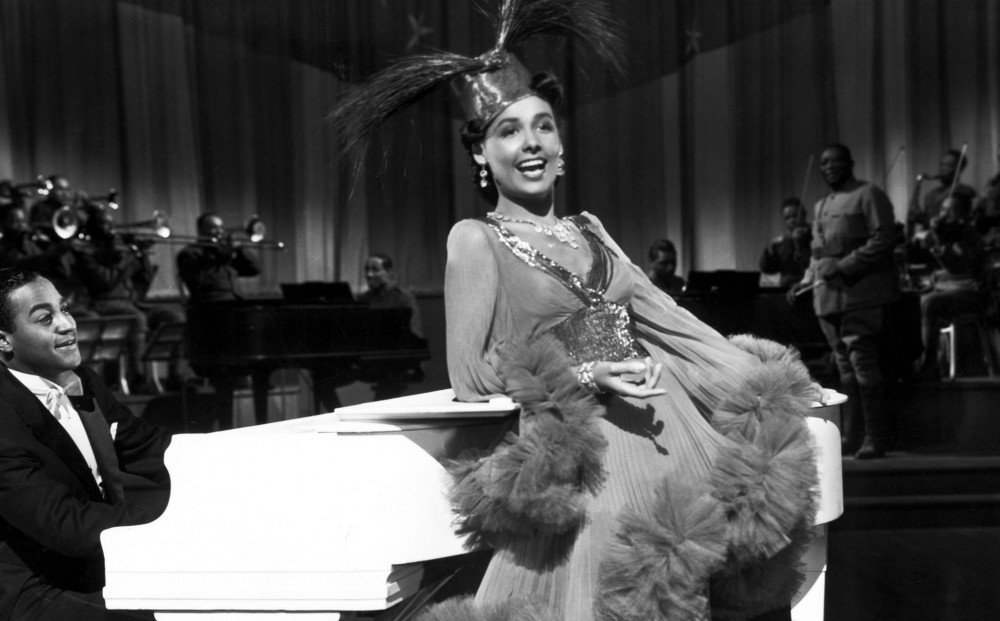 Lena Horne leans on a white piano as she sings.