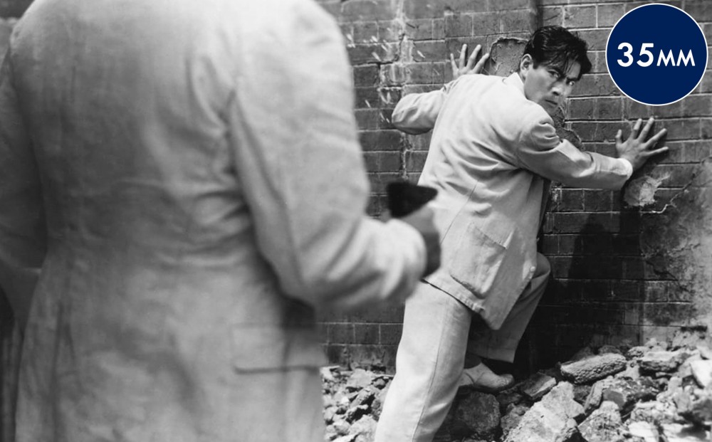 The back of a man holding a gun; he points it at a man in a white suit who has his hands on a brick wall in front of him and is looking over his shoulder.