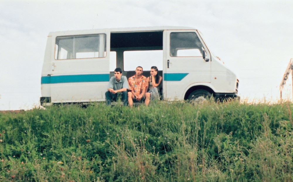 Three young people sit in their van, parked on the side of the road, looking out over a green field; the van door is slid all the way open and their feet on the roadside.