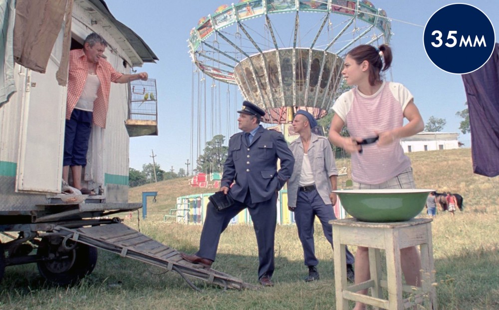 A man in uniform speaks with a man in the doorway of his trailer; a young woman stands over a bowl to prepare something while looking on, and amusement park swings are in the background.