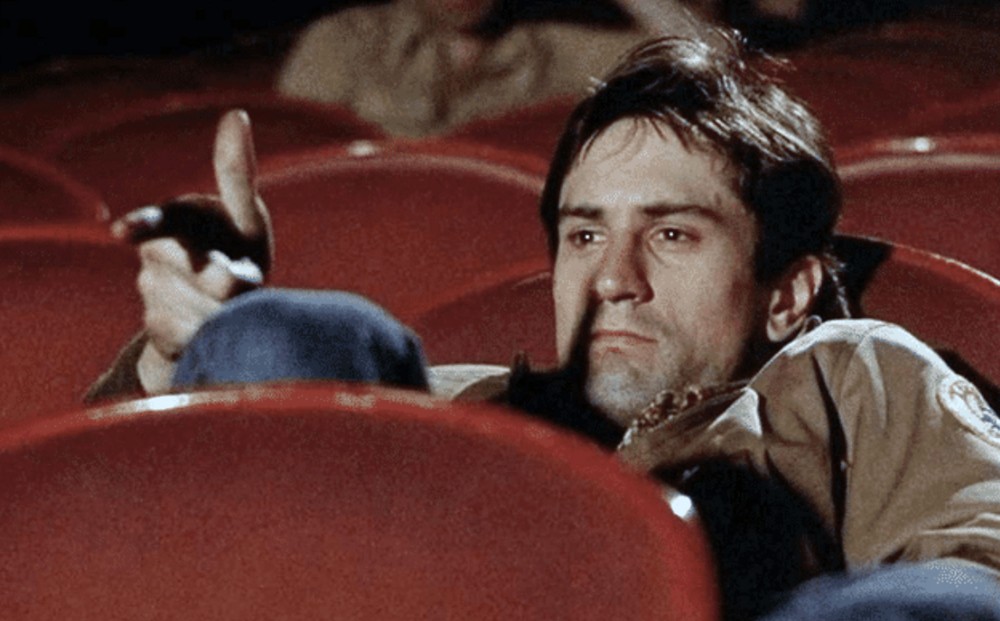 Actor Robert De Niro sits in a movie theater seat, his hand held in the shape of a gun and pointed towards the screen.