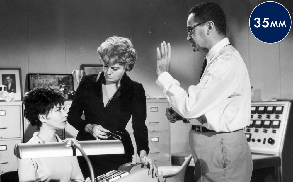 Actor Lee Grant sits at a desk, with Shelley Winters and Peter Falk standing around her.