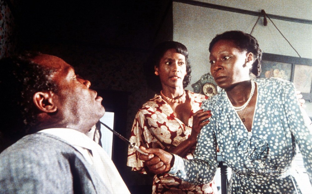 Actor Whoopi Goldberg holds a knife up to the neck of a seated Danny Glover; another woman holds Goldberg's shoulder as though to hold her back.