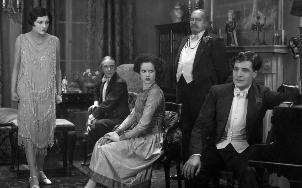 Five people in a sitting room; two women and two men look at another man (seated at a piano) with indignation.
