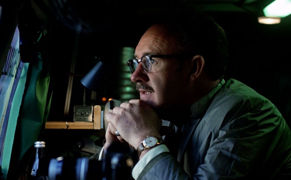 Actor Gene Hackman sits with his head rested on his hands, observing something through a window.