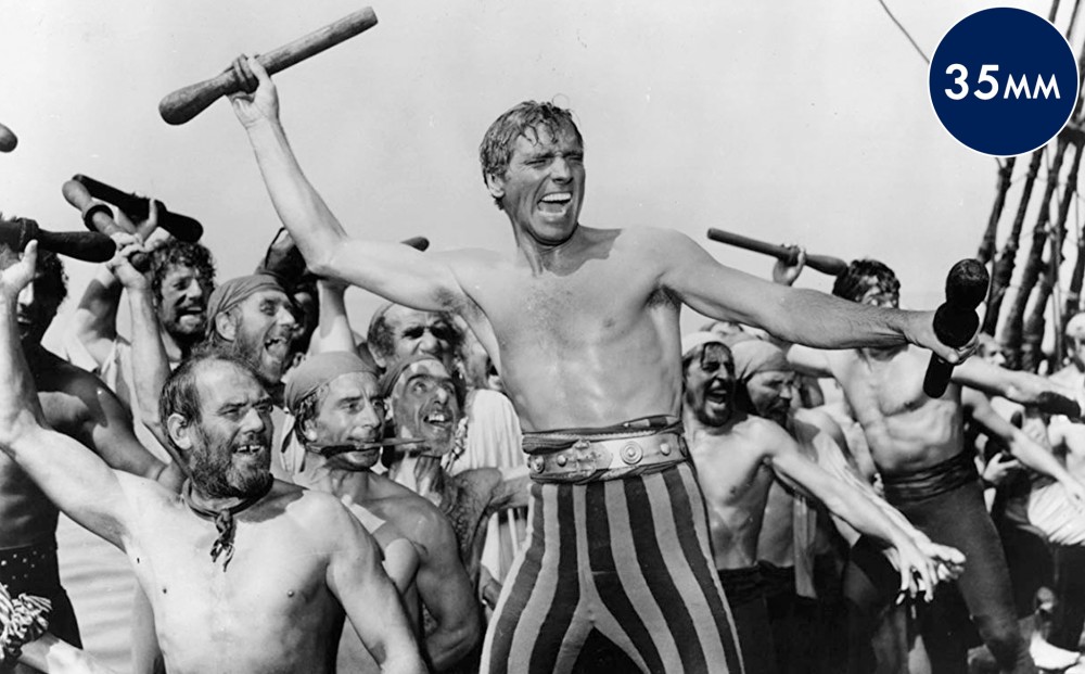 Actor Burt Lancaster brandishes two small clubs, surrounded by a motley crew of  other pirates, also wielding clubs.