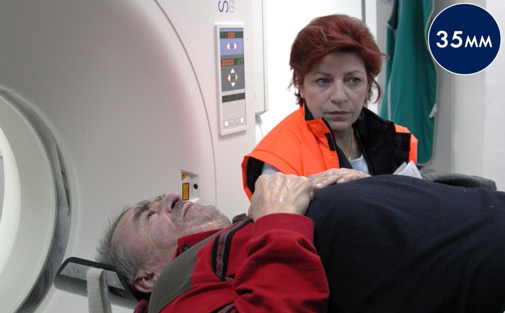 A man is laying down, ready to go into a CT scanner; a woman stands next to him.