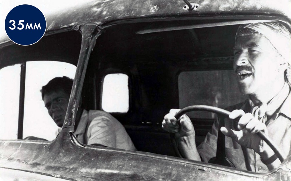 Two men sit in the front seat of a truck, one driving.