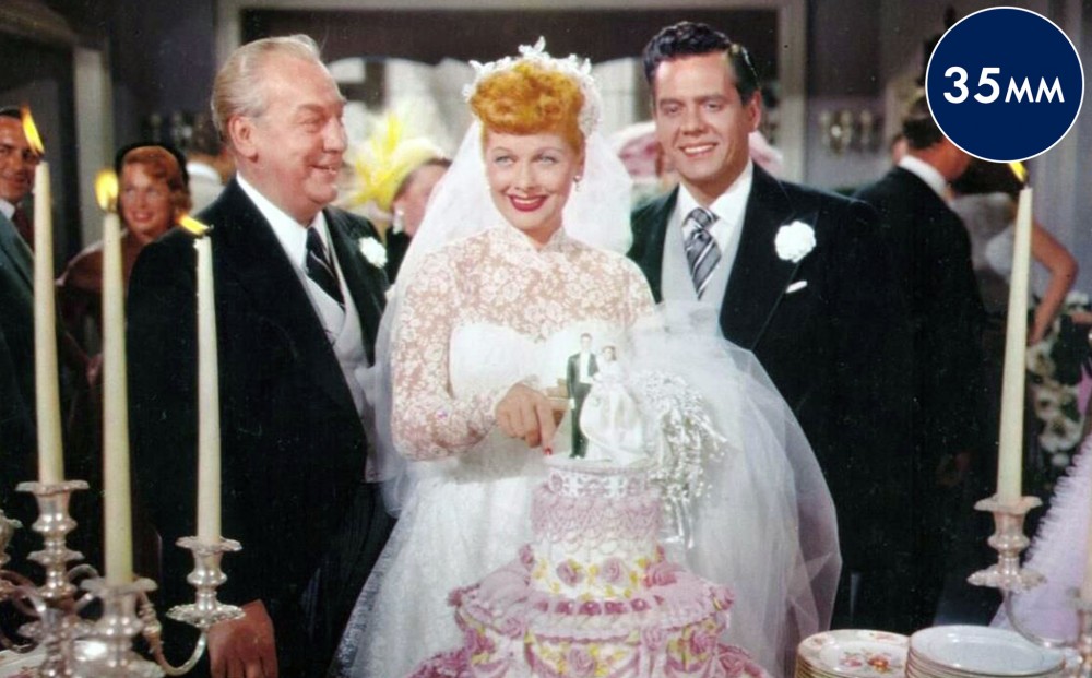 Actors Lucille Ball and Desi Arnaz smile in front of a wedding cake as bride and groom.
