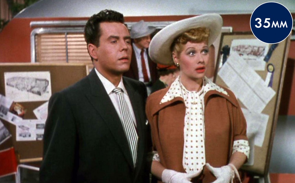 Actors Lucille Ball and Desi Arnaz stand in front of a trailer; she looks shocked.