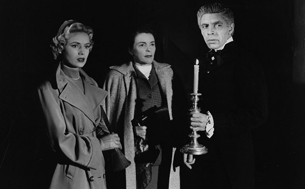 Two women stand in the dark with a man holding a tall candlestick.