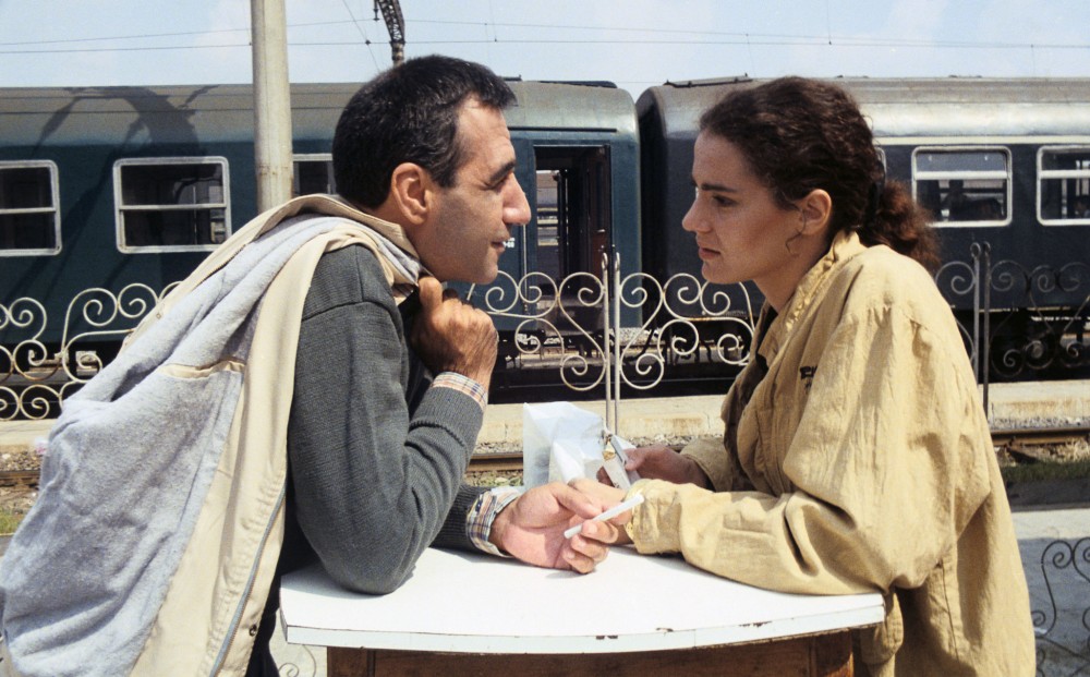 A man and woman face each other, sitting at a table in front of a train.