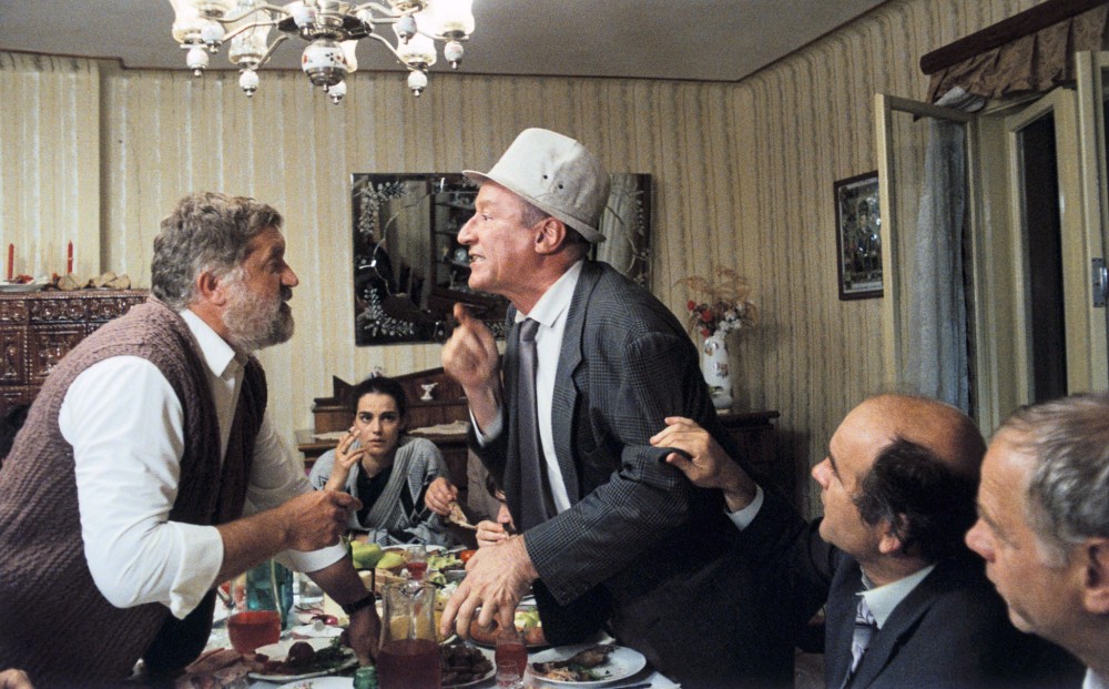 Two men are standing and arguing with each other over a dining table covered in food; others around the table try to calm them down.
