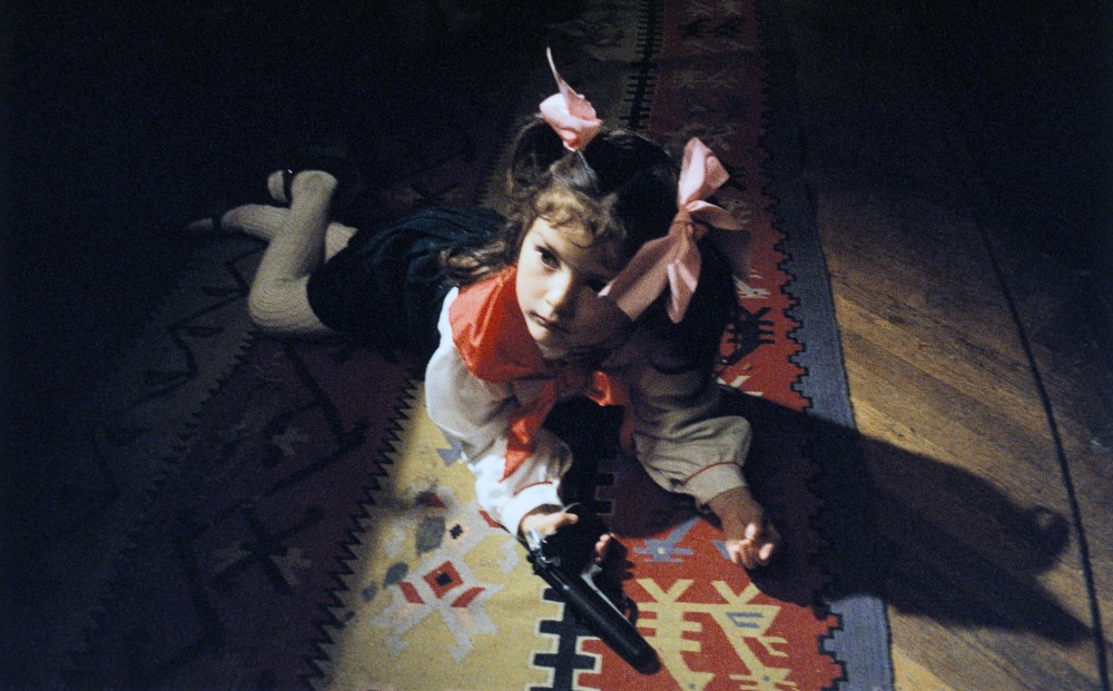 A young girl with pink bows in her hair lays on her stomach on a rug, and holds a gun.