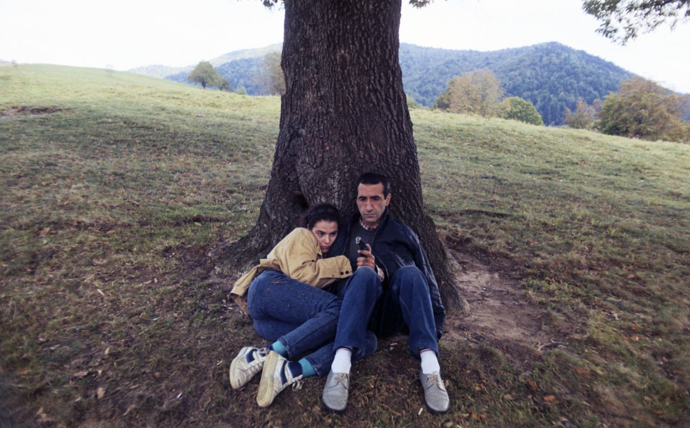 A man and woman sit together under a tree; he holds a gun.