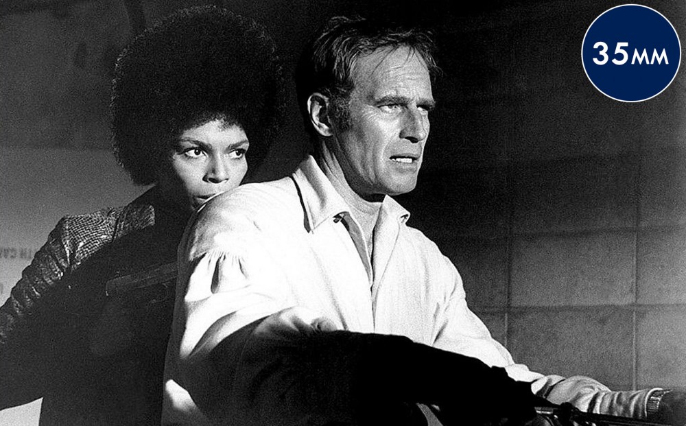Actor Rosalind Cash stands close behind Charlton Heston; she holds a gun to his back.