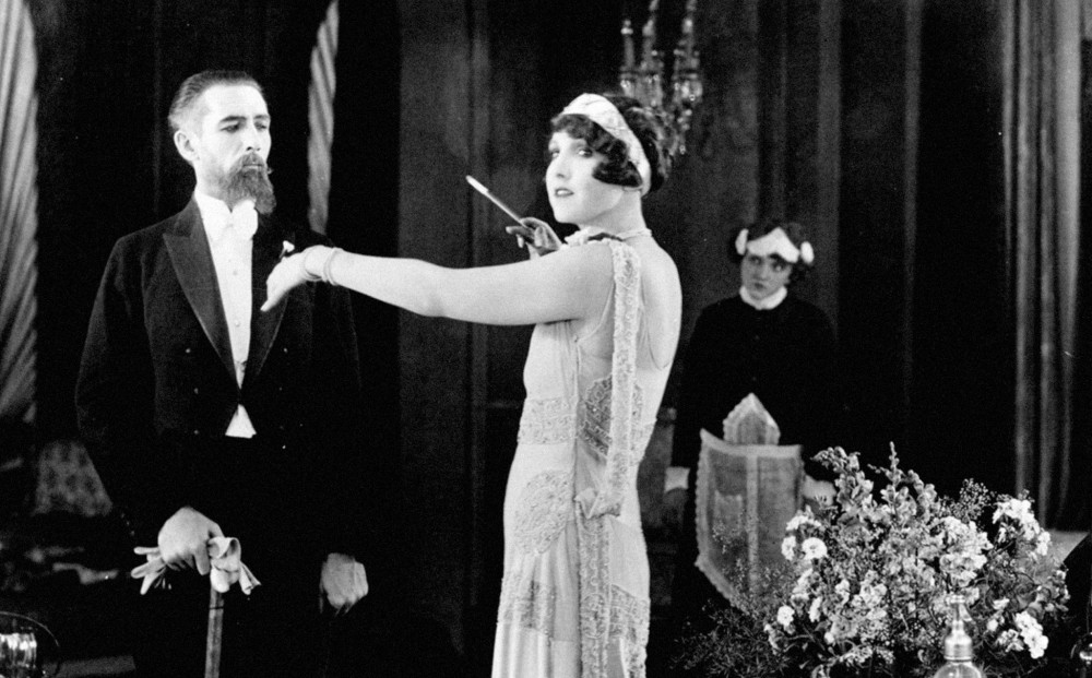 A woman dressed in a long evening gown stands next to a man in a coat and tails; a servant hovers in the background.