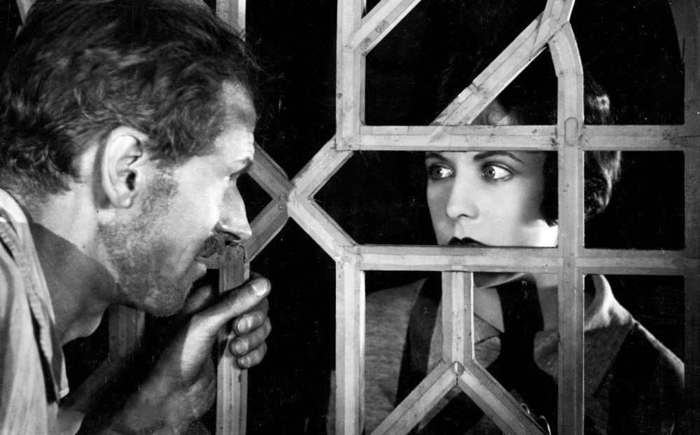 A man and a woman look at each other through a lattice.