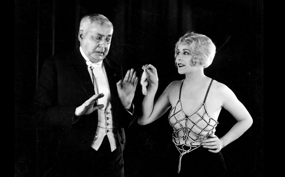 A woman holds out a piece of curly hair towards a man; he holds his hands up and looks nervous.