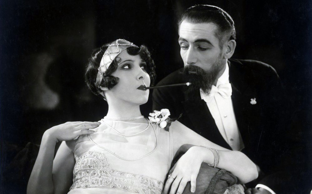 A woman lounges with a long cigarette in her mouth; a man sits next to her.