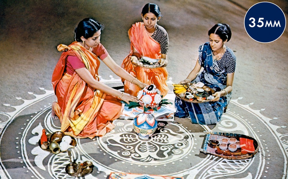 Three women in saris sit on the floor with a few platters of food.