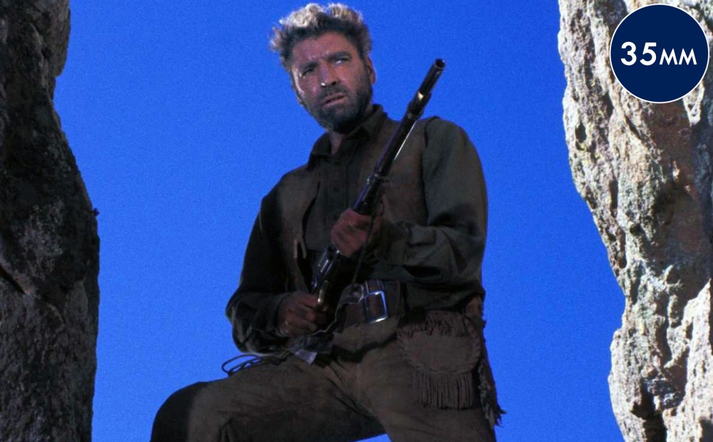 Actor Burt Lancaster stands between two rocks, holding a gun.