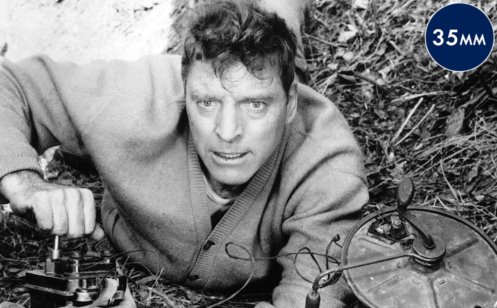 Actor Burt Lancaster lays on his stomach on the ground, working with tools.