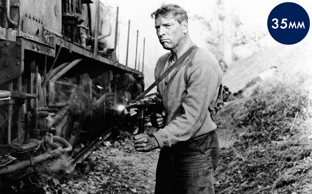 Actor Burt Lancaster holds a machine gun, standing next to a train.