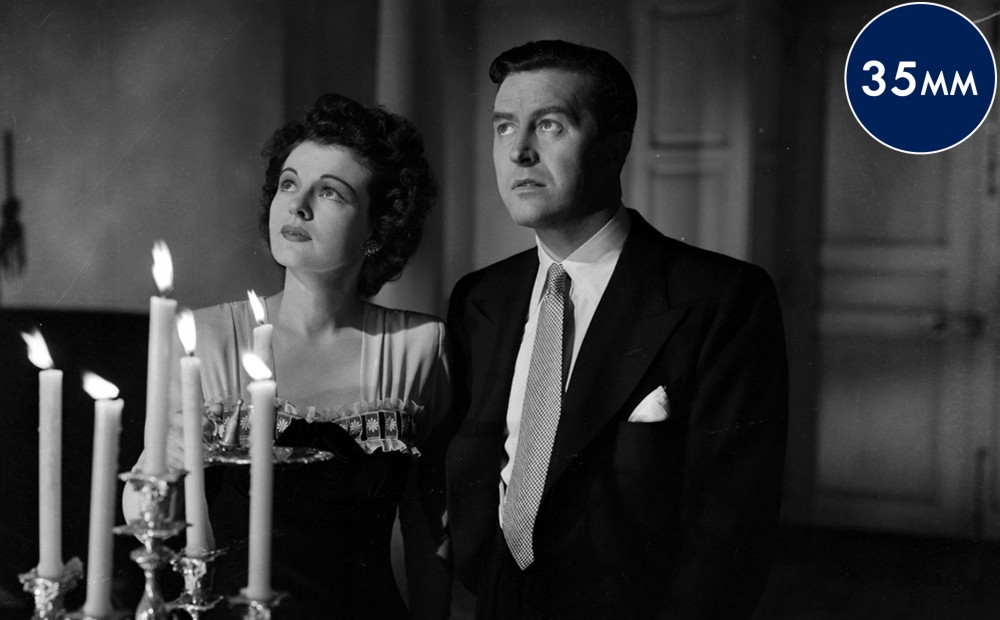 Actors Ray Milland and Ruth Hussey look upwards; long candlesticks in front of them.