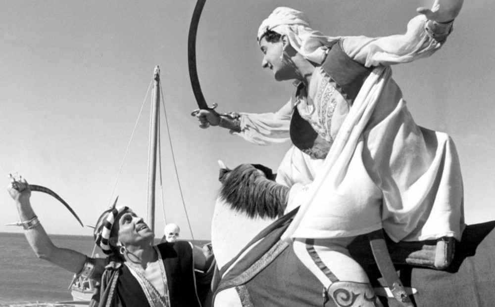 The white sheik swings his sword at a man who is dressed like a pirate; the sheik is on a horse.