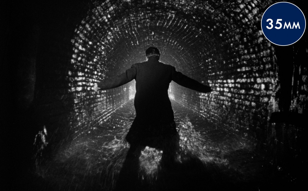 A man in a long coat stands in a tunnel, his back to the camera.