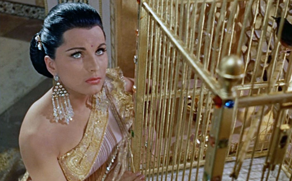 From THE TIGER OF ESCHNAPUR: Actor Debra Paget looks up, kneeling beside a gold bird cage.